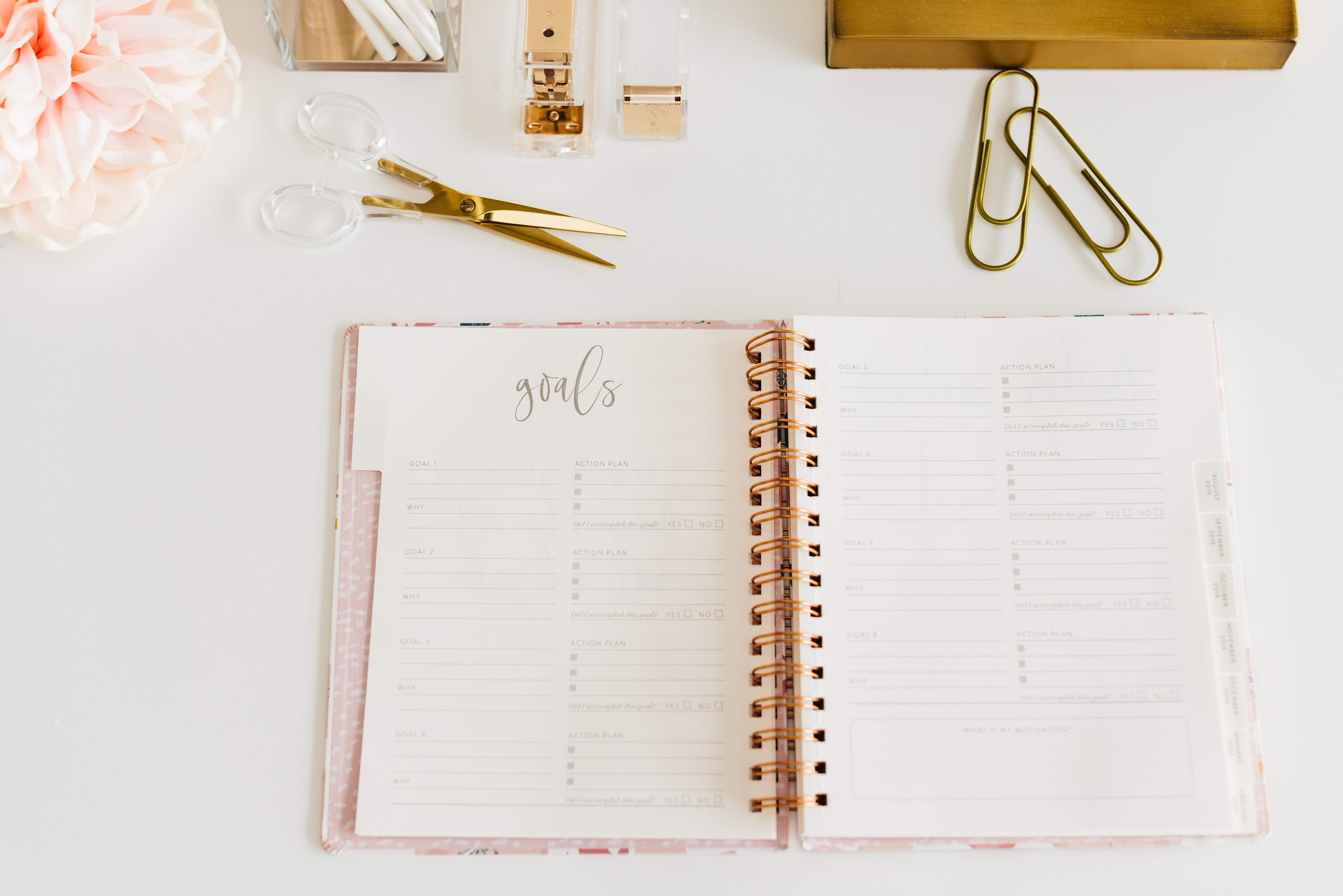 Journal open on a table with paper clips and a scissors and pretty flowers around it. The notebook has a heading saying goals.